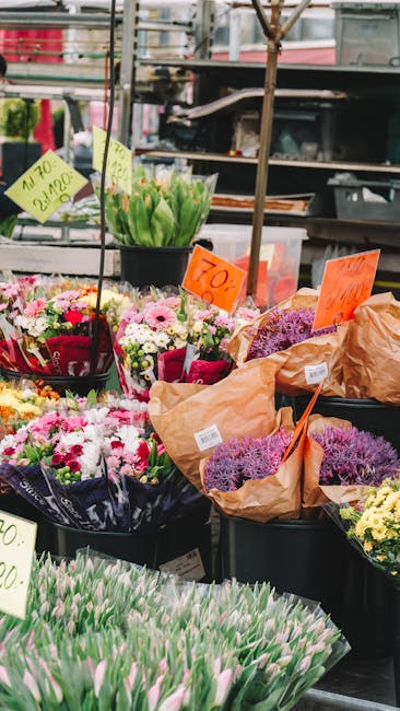 A vibrant outdoor flower market display featuring an assortment of fresh floral bouquets and individual blooms. Prominent in the foreground are wrapped tulip bundles with pink and white petals, arrang