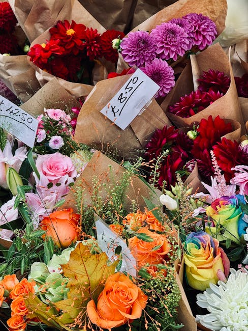 A vibrant assortment of freshly cut flowers displayed in brown paper wrapping at a floral market, featuring bright pink dahlias, deep red chrysanthemums, orange roses, pale pink lisianthus, and rainbo