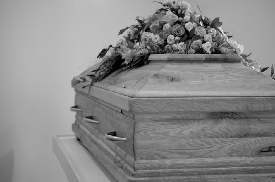 A wooden casket with a smooth, polished surface and visible natural wood grain, resting on a white table against a plain background. The casket is draped with an elegant floral arrangement consisting 