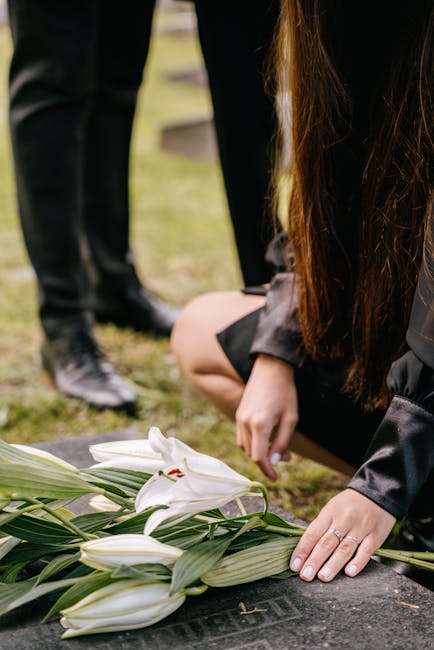 A person laying a bouquet of white lilies with lush green leaves on a gravestone during a funeral, with another individual standing nearby in the background. The lilies are fresh, with smooth petals a