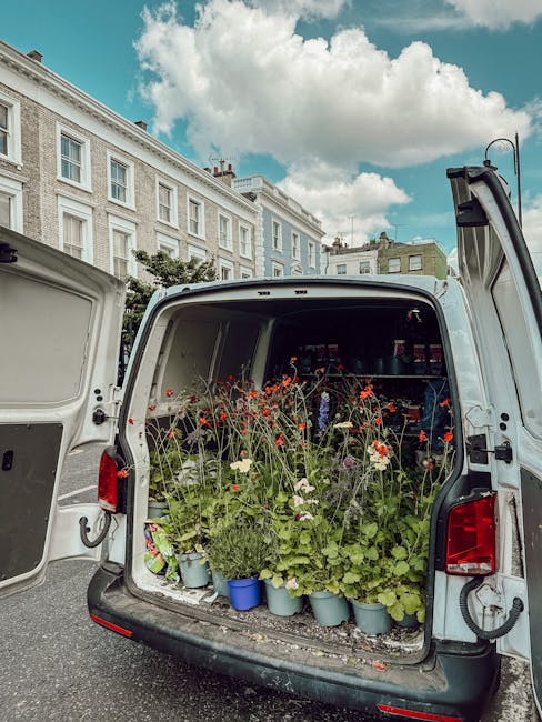 The back of a white delivery van opened to reveal an interior filled with freshly picked flowers and green foliage, including red poppies, white daisies, and various leafy stems, arranged in multiple 