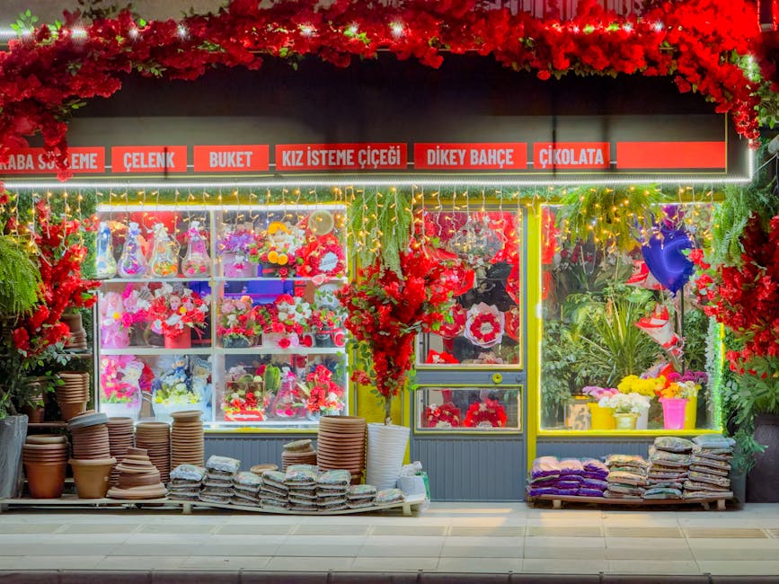 The image depicts a flower shop storefront with a vibrant display of fresh floral arrangements and potted plants. The shop is decorated with red flower garlands and illuminated with warm, yellow strin