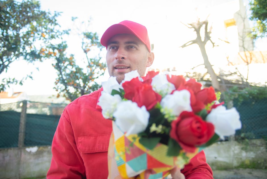 A male delivery person dressed in a red uniform and matching red cap holds a vibrant bouquet of fresh flowers, including red roses, white lisianthus, and green foliage, wrapped in colorful paper. He i