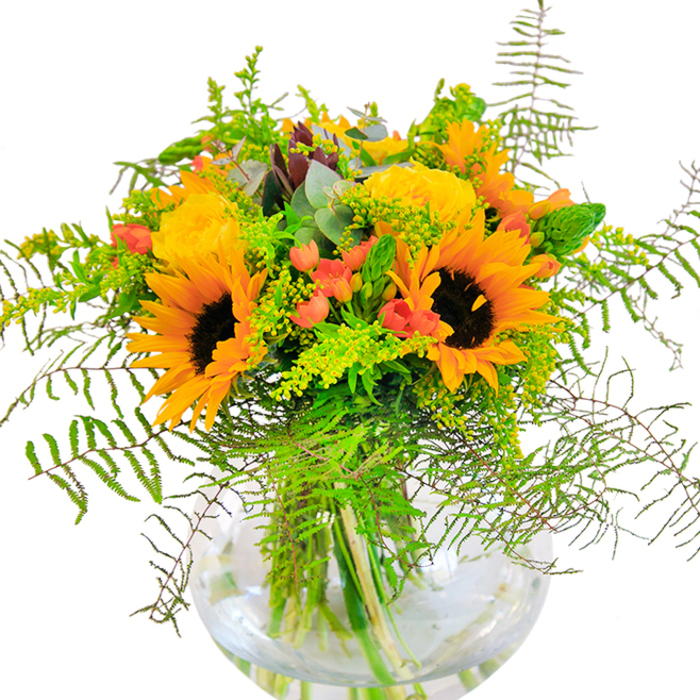 The first impression of this magnificent Brent bouquet is a celebration of sunlight and city gardens, with vibrant sunflowers and densely layered yellow roses arranged in a modern, round glass vase. Each sunflower's dark center is ringed with bright, inviting yellow-orange petals-reminiscent of the golden afternoons in Kensal Green or the playful vibrance of Willesden's summer fairs. Interspersed are garden roses, their ruffled petals glowing like buttery clouds, imparting elegance amid the free-spirited array of foliage. Ruby red Hypericum berries sparkle among sprigs of fresh greenery, echoing berries seen in Brent's hedgerows and masterfully drawing the eye. The bouquet's lushness is heightened by delicate, trailing ferns and slender spikes of green filler, their differing textures suggesting both wild abundance and precise florist craftsmanship. Pale yellow solidago and rounded eucalyptus leaves complete the composition, adding subtle softness and a breath of fresh air-details that bring to mind early mornings by the Regent's Canal. The clear vase grounds the arrangement, revealing the clean alignment of stems and water that amplify its sense of freshness. Each element-color, form, and scent-comes together to create a visual and sensory celebration of Brent life. Designed for milestone celebrations, heartfelt thank-yous, or simply to bring a smile, this arrangement translates everyday beauty into a living work of art.